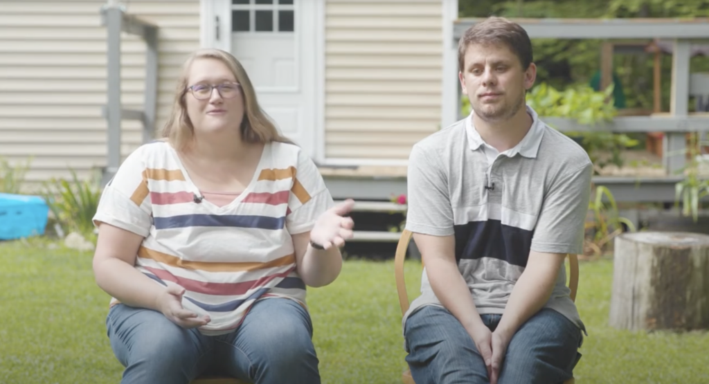 Two people sit outside in front of a beige house. The person on the left gestures with one hand while speaking; both are seated on chairs on the grass.