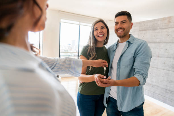 A woman hands keys to a smiling couple standing together in a bright, modern apartment, suggesting they are receiving or renting the property.