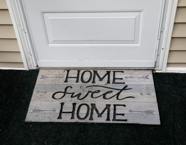 A doormat with the words "Home Sweet Home" is placed in front of a closed white door on a dark outdoor carpet.