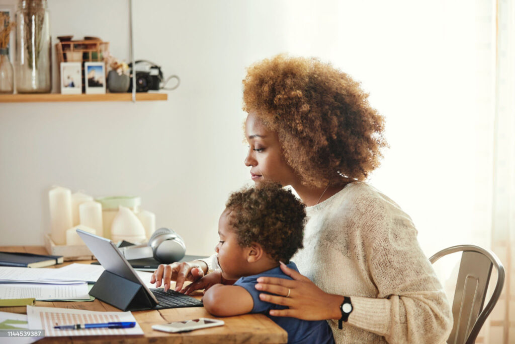 A woman works on a laptop at a wooden table while a young child sits on her lap, both looking at the screen. Various office supplies and books are on the table.
