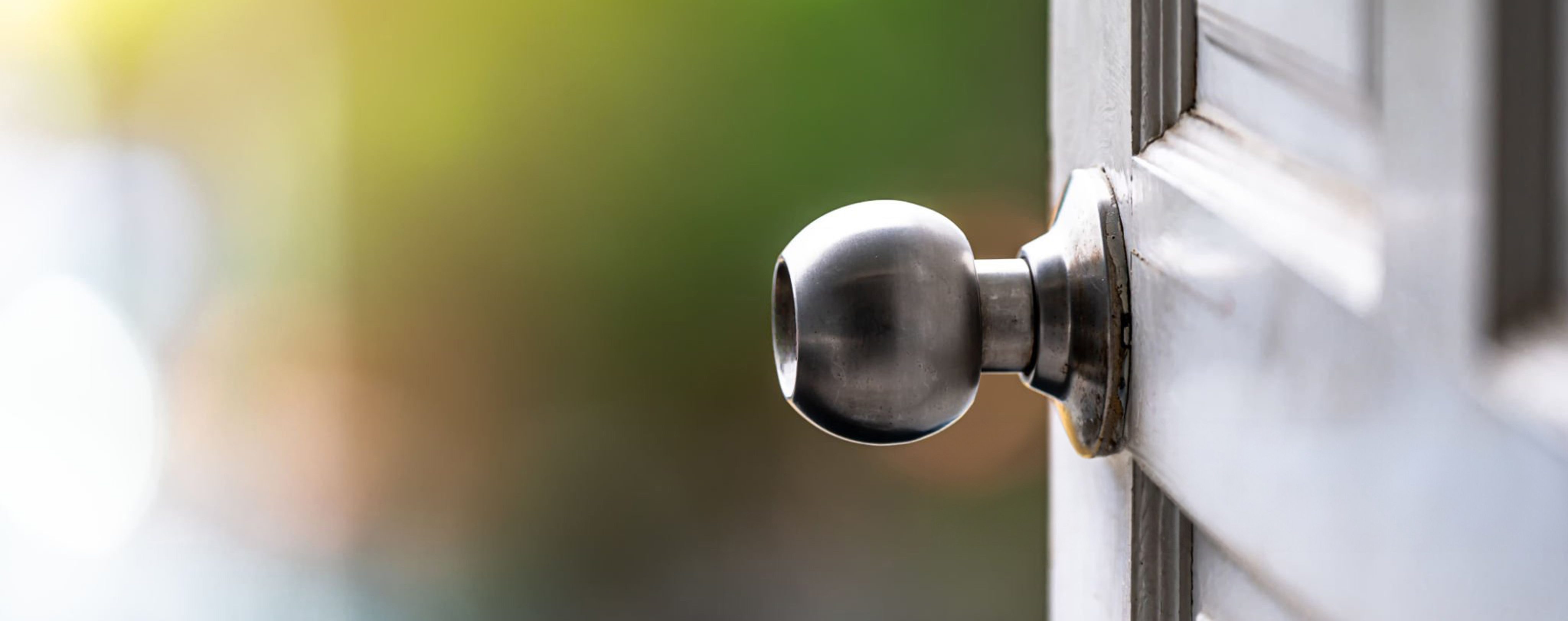 A close-up of a round silver doorknob on a partially open white door, with a blurred green and yellow background outside.