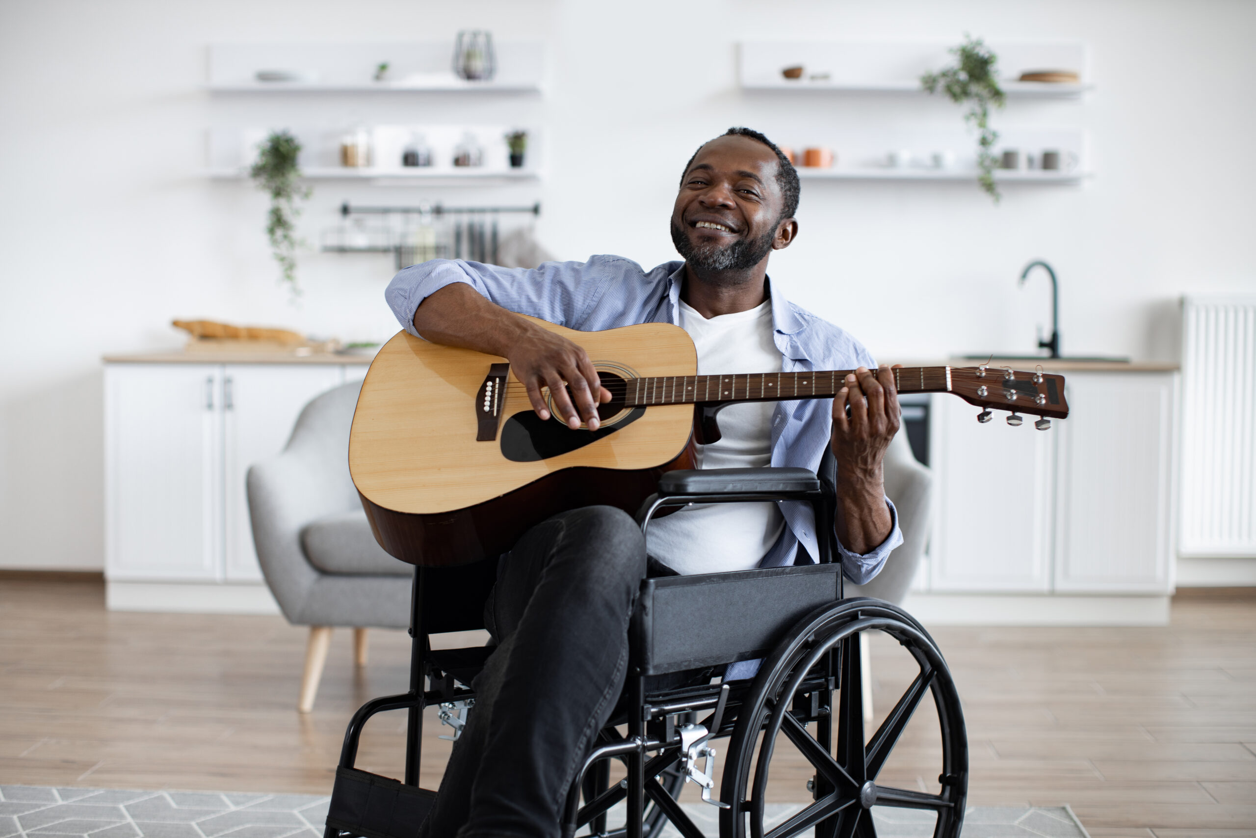 A man sitting in a wheelchair plays an acoustic guitar and smiles in a modern, bright living room.