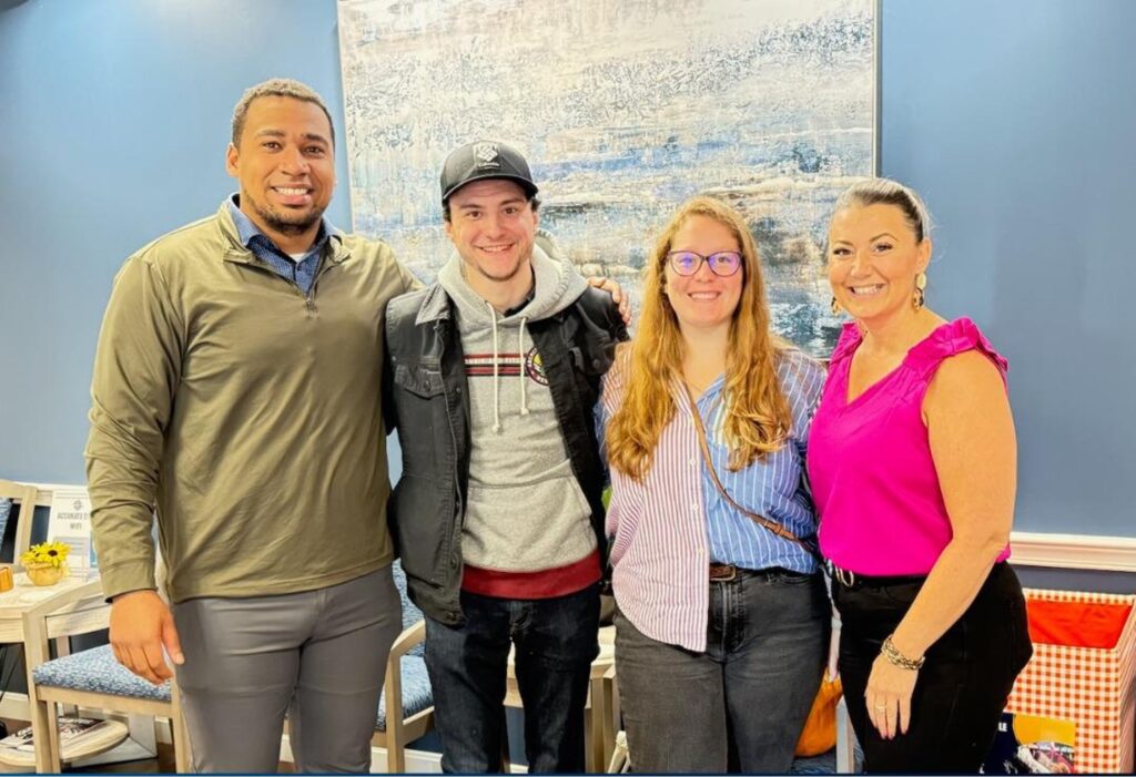 Four people stand side by side indoors, smiling at the camera. A blue abstract painting hangs on the wall behind them.