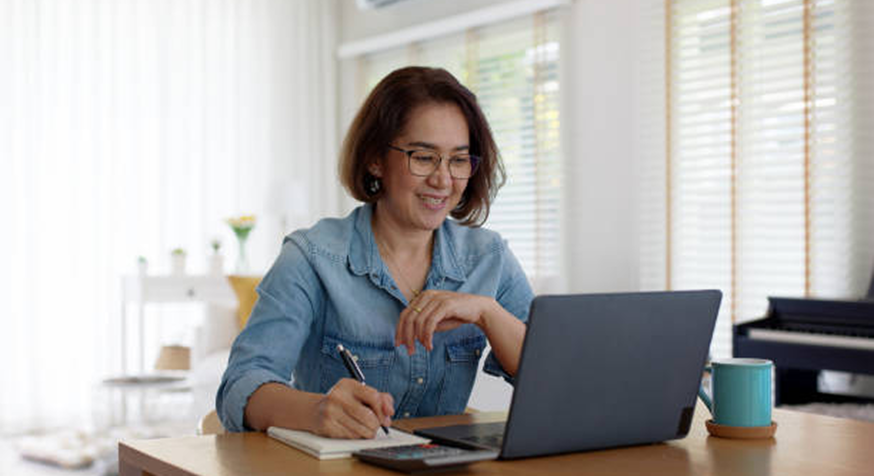 A woman wearing glasses works at a laptop, taking notes in a notebook with a pen. A calculator, mug, and other items are on the desk in a bright room.