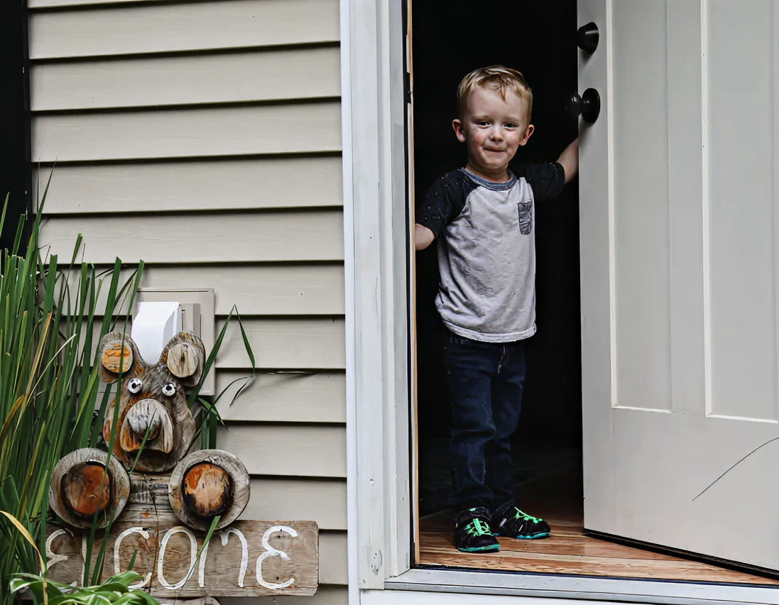 A young boy stands smiling in the doorway of a house next to a wooden welcome sign with a bear decoration.