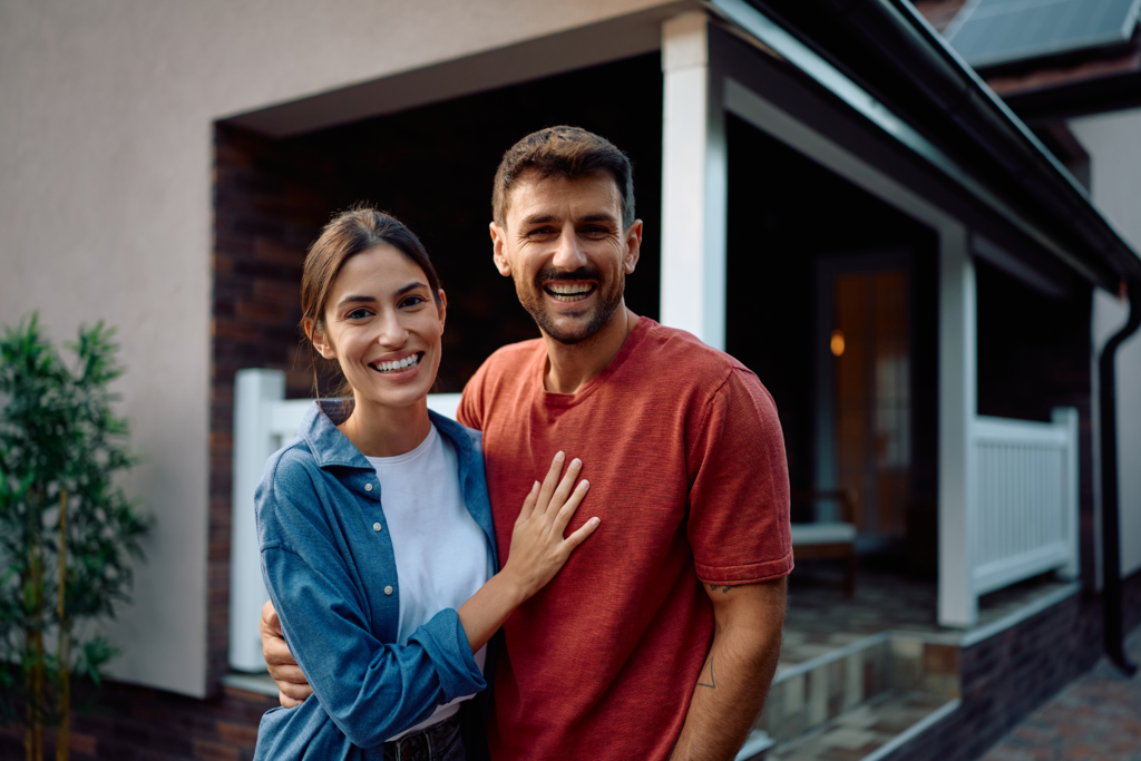 A smiling couple stands close together in front of a house, posing for the camera.