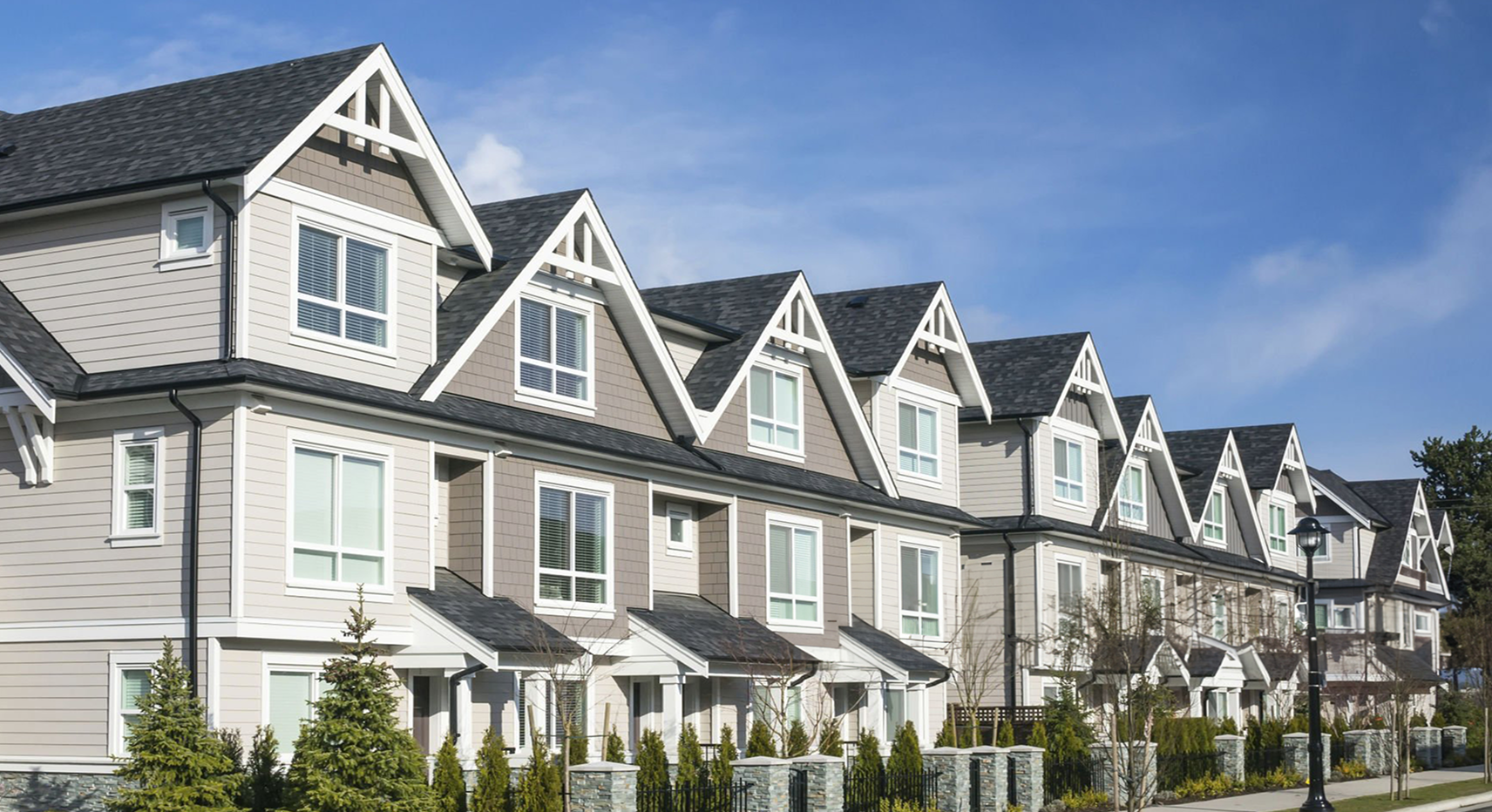 Row of modern, two-story townhouses with gabled roofs, large windows, and small landscaped front yards under a blue sky.