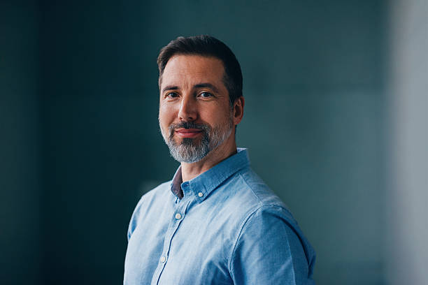 A man with short dark hair and a trimmed gray beard is wearing a light blue button-up shirt and smiling slightly, standing against a plain dark background.