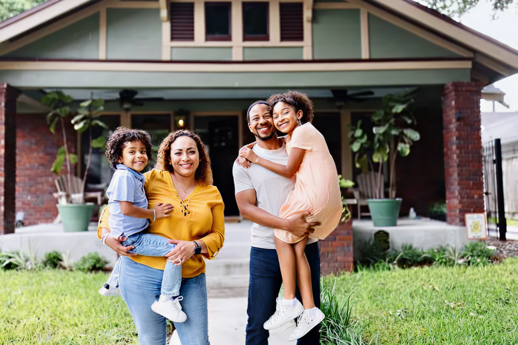 A family of four stands smiling in front of a house; two adults each hold a child in their arms on a grassy lawn.
