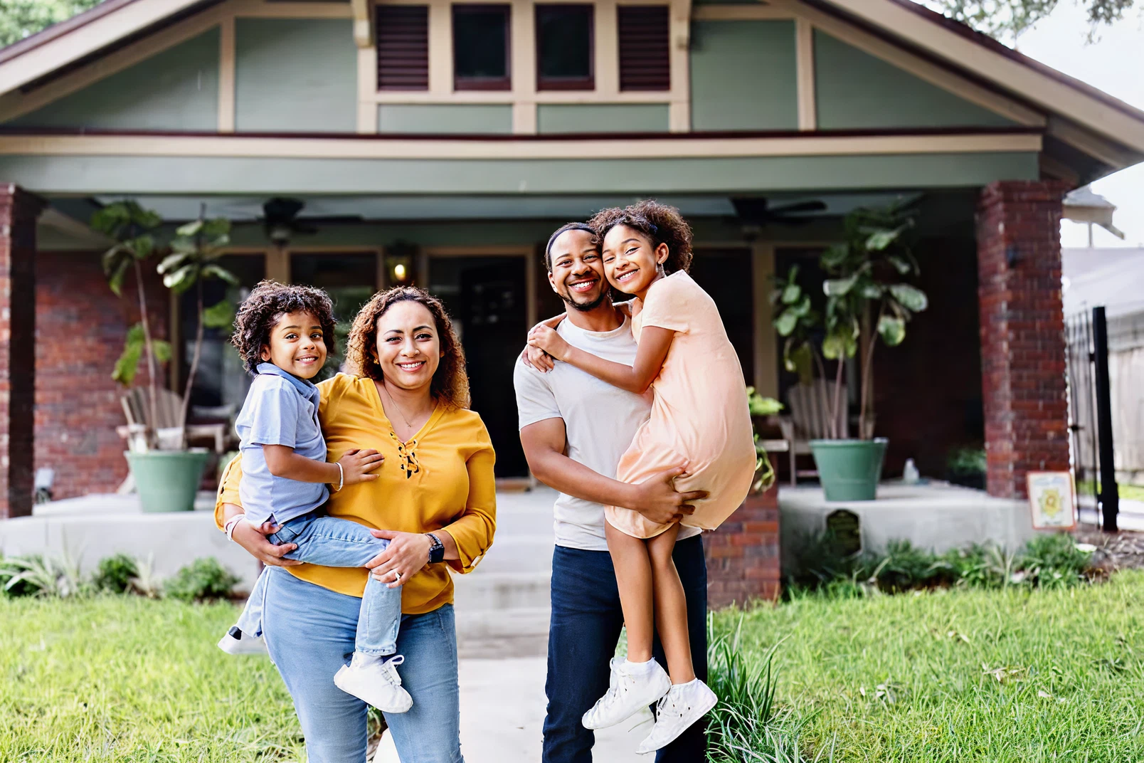 A family of four stands smiling in front of a house; two adults each hold a child in their arms on a grassy lawn.
