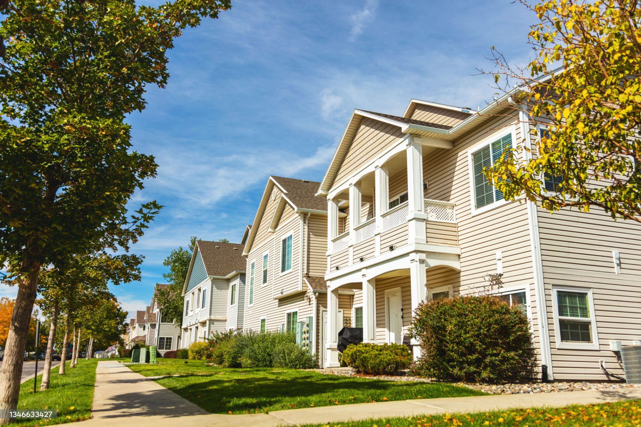 Row of modern suburban houses with light-colored siding, front porches, and neatly maintained lawns along a tree-lined sidewalk under a blue sky.