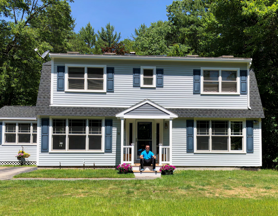 A two-story blue house with white trim and blue shutters sits on a lawn. A person is seated on the front steps near potted flowers, with trees in the background.