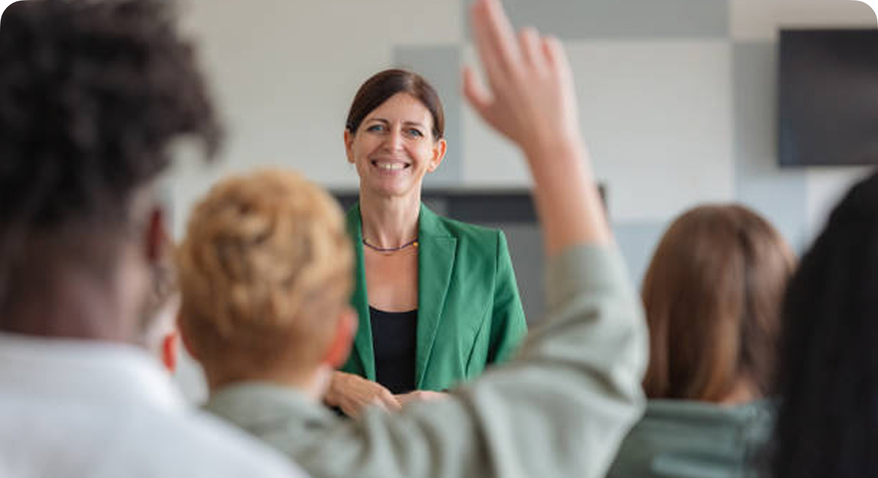 A woman stands at the front of a classroom smiling, while a student raises their hand to ask a question. Other students are seated and facing her.