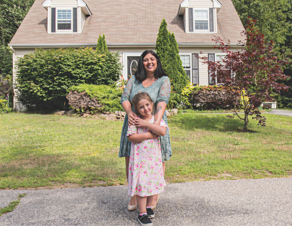 Adult woman and young girl standing and smiling in front of a suburban house with trees and grass in the background.