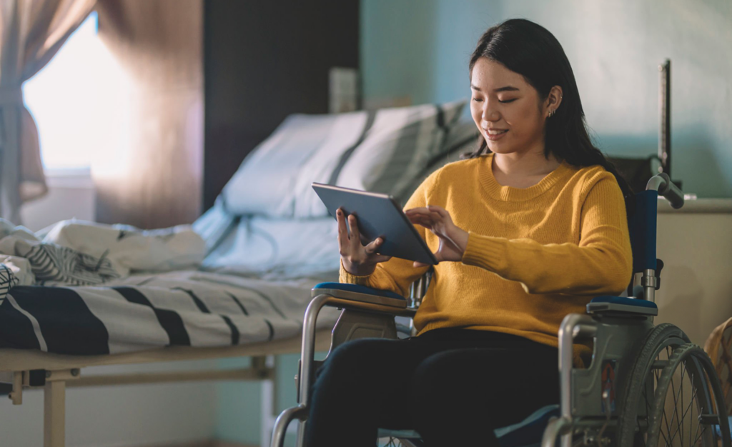 A woman in a wheelchair uses a tablet while sitting in a bedroom with an unmade bed and natural light coming through a window.
