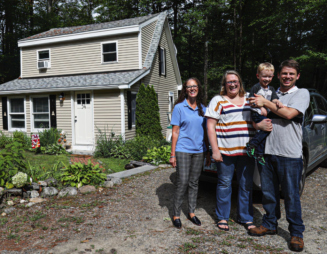 Four people stand and smile in front of a tan house with a garden. One adult holds a young child. A car is parked next to them. Trees are visible in the background.
