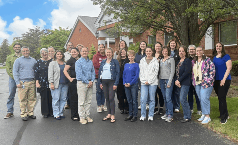 A group of 22 adults stand outside a red brick building on a paved walkway, posing for a group photo under a tree.