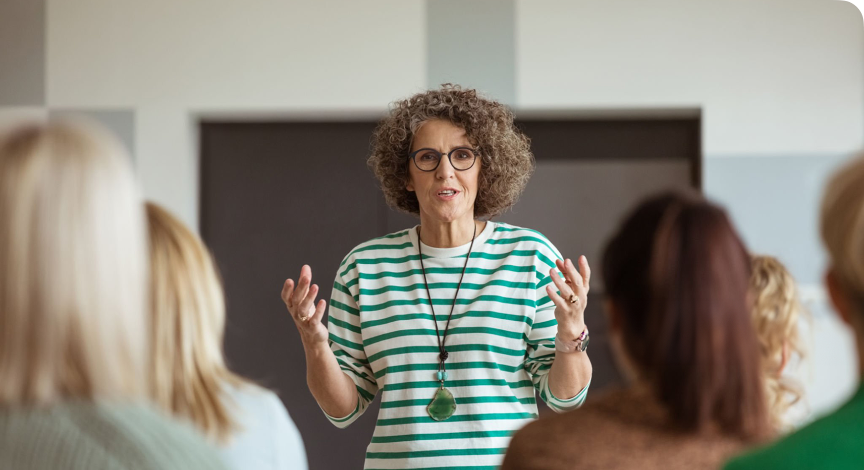 A woman with curly hair and glasses speaks to a seated group, gesturing with her hands, in a modern indoor setting.