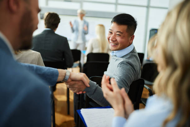 Man smiling and shaking hands with another person while sitting in an audience at a seminar or meeting, with people and a speaker visible in the background.