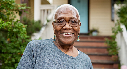 An older woman with short gray hair and glasses smiles while standing outside in front of a house with stairs and greenery.