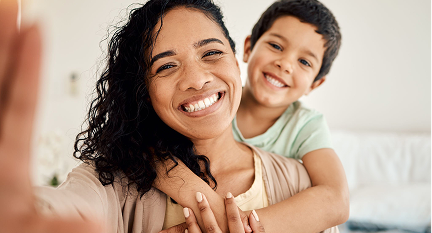 A woman smiling and taking a selfie with a young boy who is hugging her from behind, both looking happy indoors.