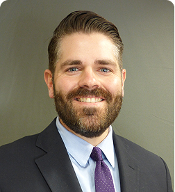 Man with a beard and mustache wearing a dark suit, light blue shirt, and purple tie, smiling in front of a plain gray background.