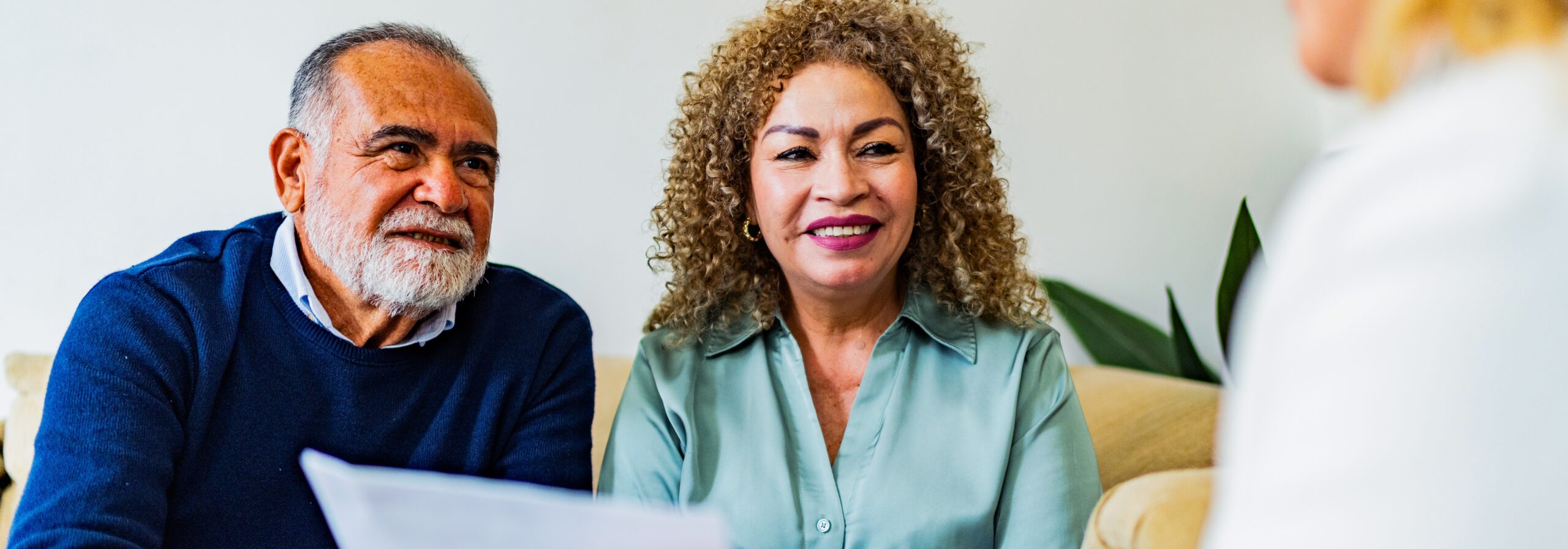 An older man and woman sit on a couch, attentively listening to a person holding papers in front of them.