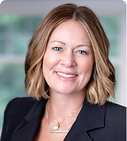 Woman with shoulder-length blond hair wearing a black blazer and necklace, smiling, with a blurred background.