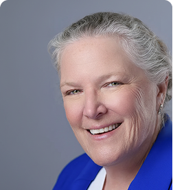 Smiling older woman with gray hair pulled back, wearing a blue blazer and white top, against a plain light gray background.