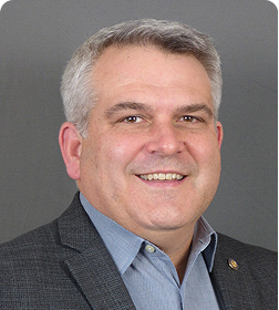 A smiling middle-aged man with short gray hair wearing a gray suit jacket and light blue shirt, posed against a plain gray background.