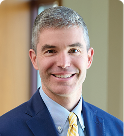 A man with short gray hair in a blue suit, light blue shirt, and yellow tie, smiling in an indoor office setting.