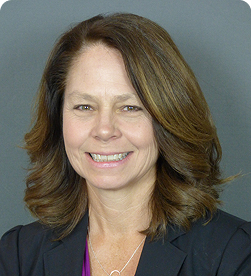 Smiling woman with shoulder-length brown hair wearing a dark blazer over a purple top, posed in front of a plain gray background.