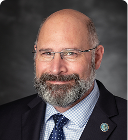 A middle-aged man with a bald head, gray beard, and glasses, wearing a suit, blue checkered shirt, polka dot tie, and a lapel pin, poses in front of a dark background.