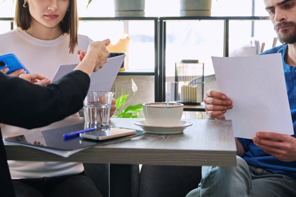 Three people sit at a table with documents, a smartphone, a pen, a glass of water, and a cup of coffee in front of them, appearing to discuss or review paperwork.