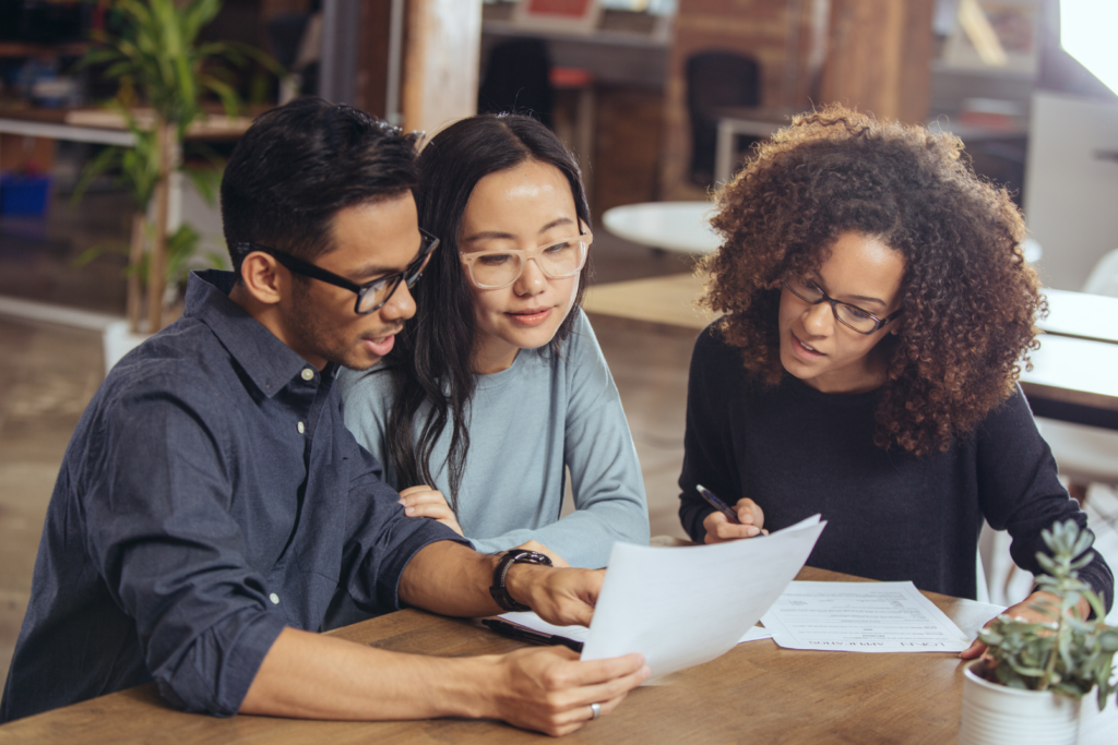 Three people sit at a table, looking at documents together, discussing and reviewing papers in a modern office setting.