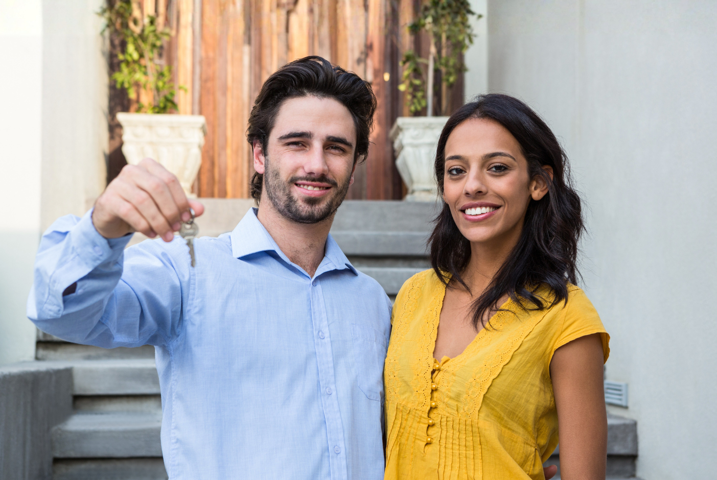 A man and a woman stand outside a house, smiling, while the man holds up a set of keys.