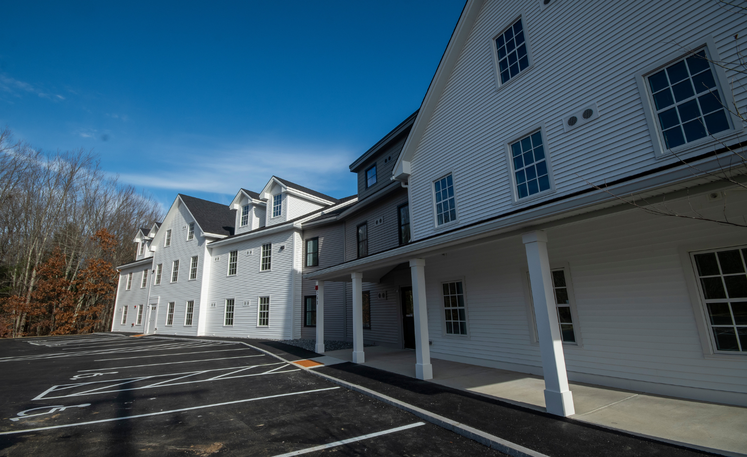 A white, multi-story residential building with a covered entrance and an empty parking lot under a clear blue sky.