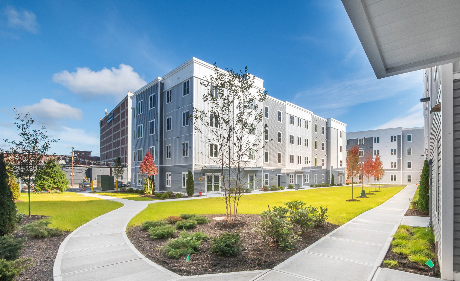Modern apartment complex with light gray buildings, landscaped green lawns, young trees, and walkways under a blue sky.