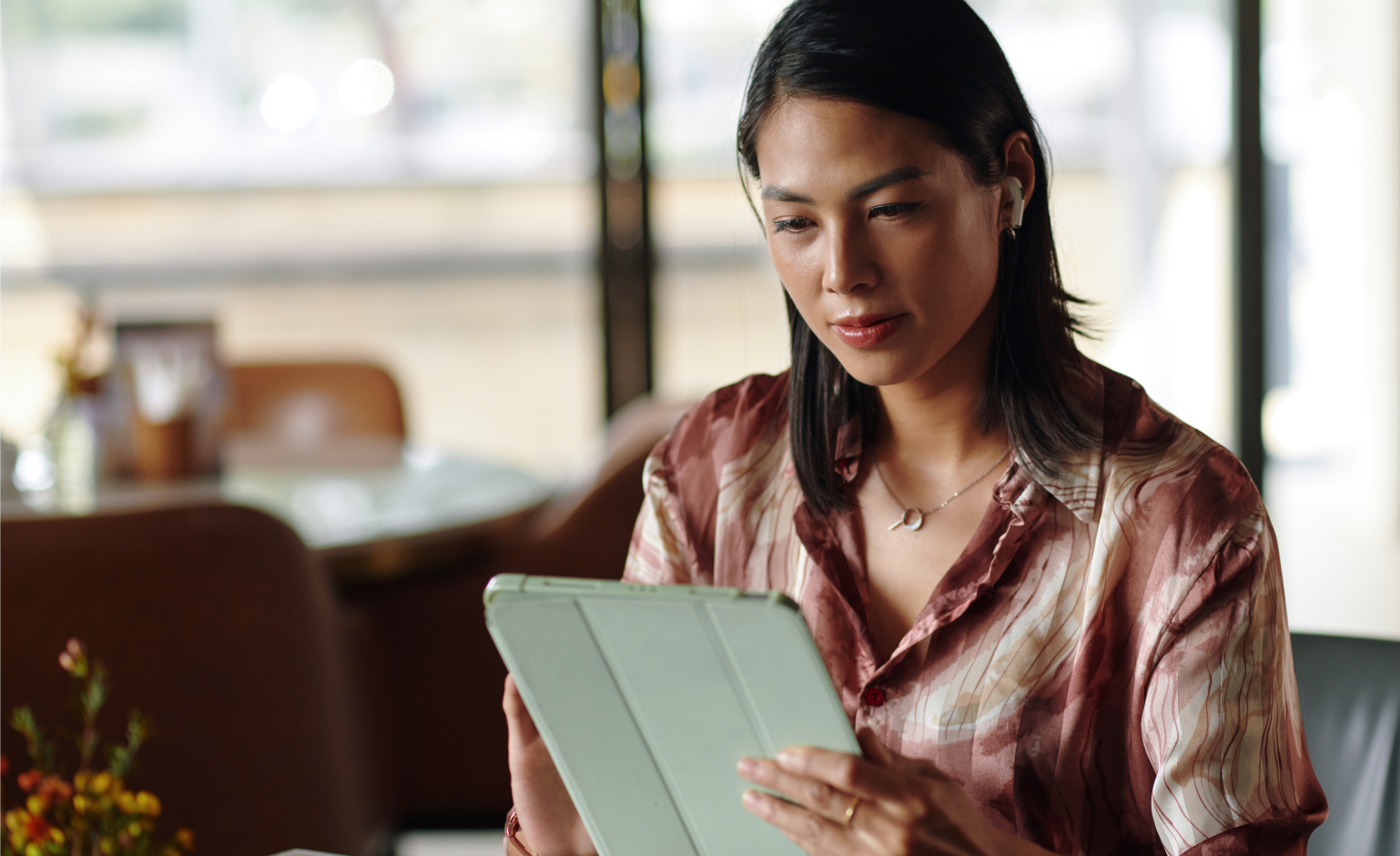 A woman sits at a table in a cafe, looking at a tablet. She is wearing a patterned blouse and has straight shoulder-length hair.
