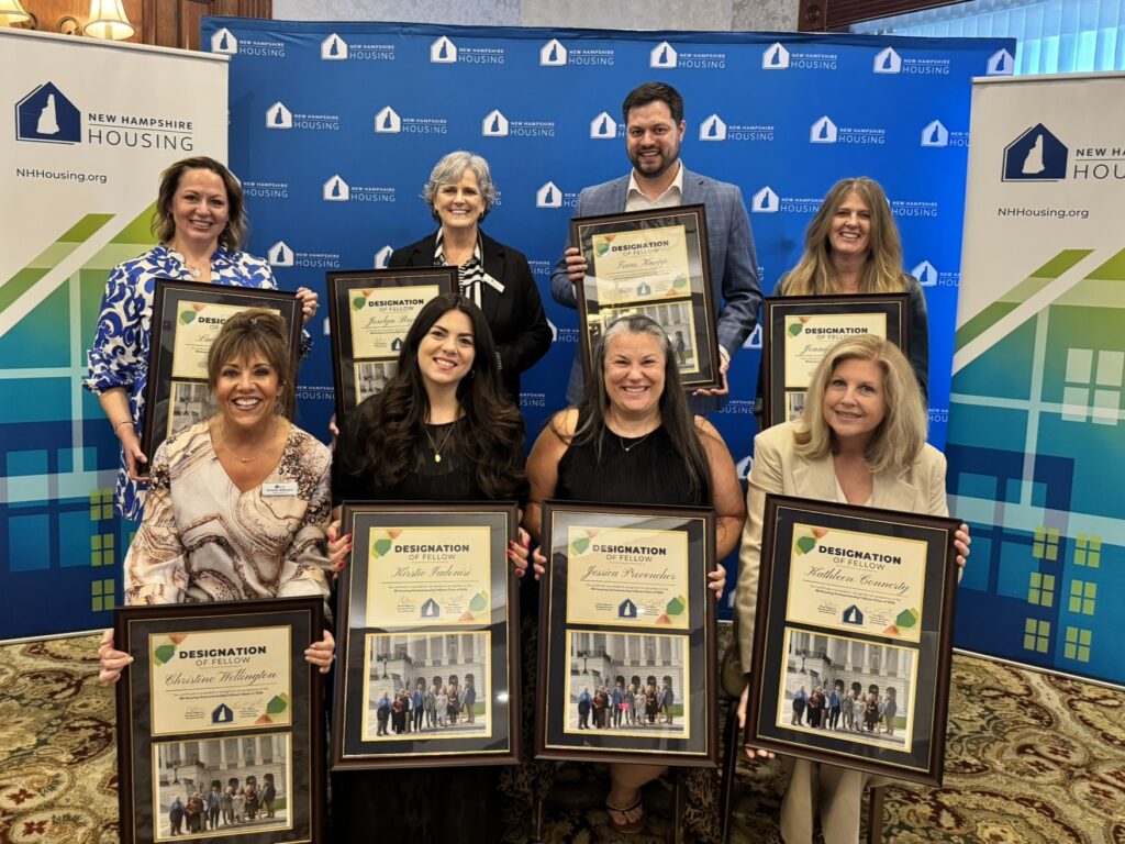 Eight people pose indoors holding framed awards in front of New Hampshire Housing banners, smiling for a group photo.