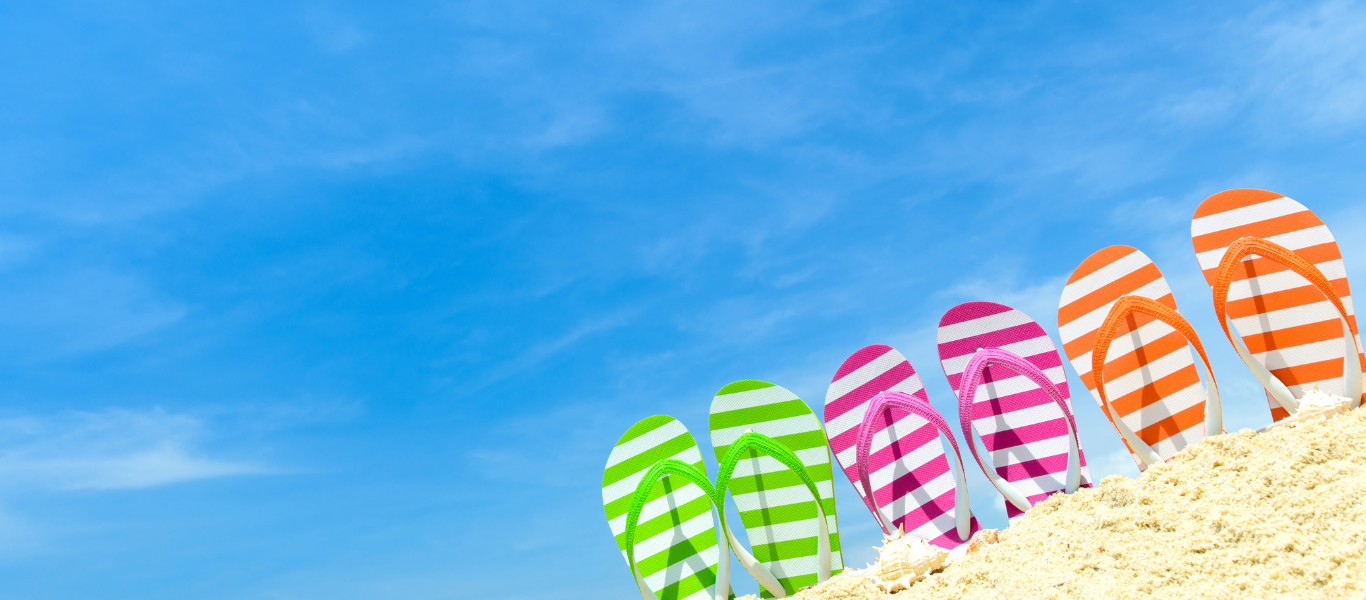 Six pairs of colorful striped flip-flops are lined up in sand under a blue sky.