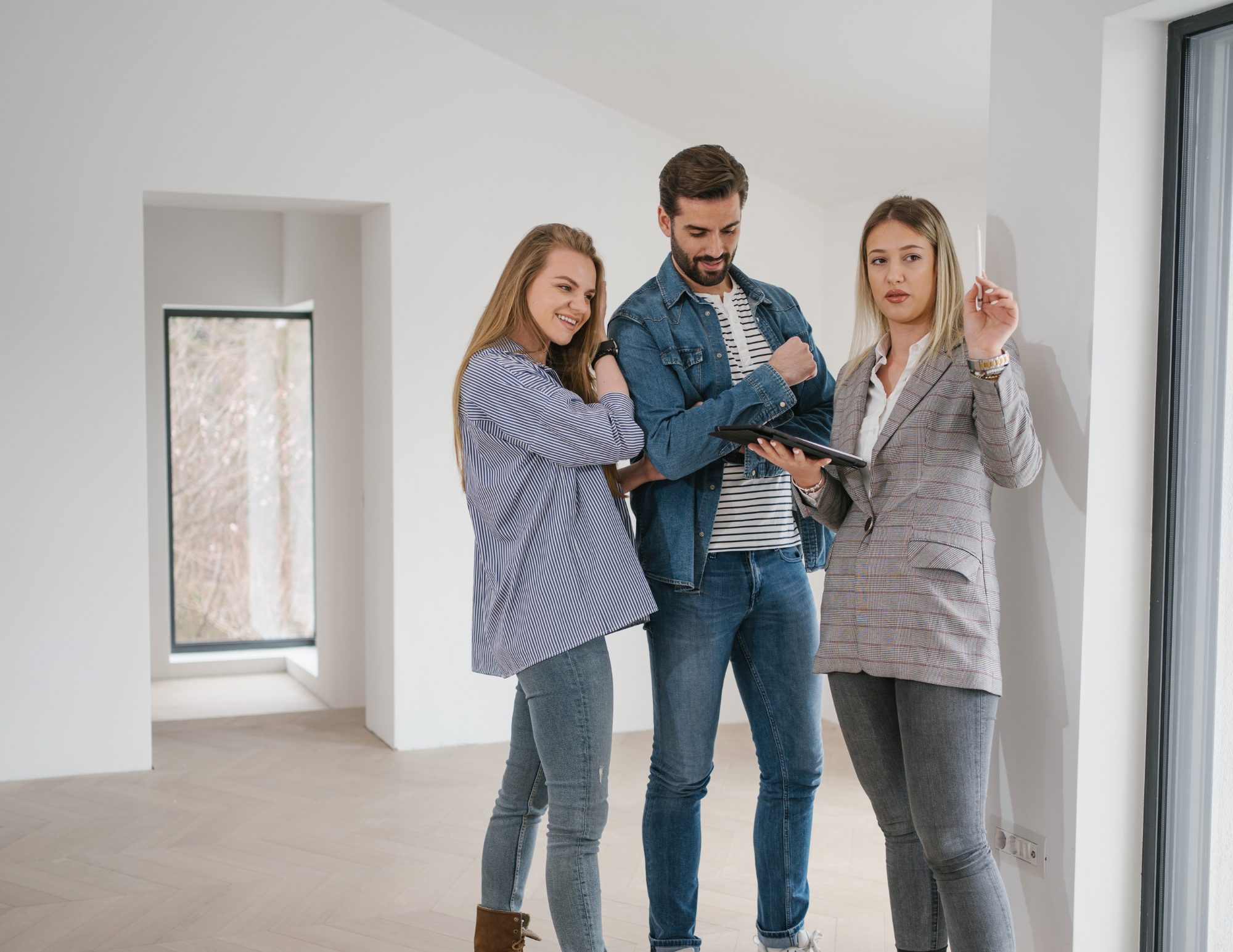 Three people stand in a bright, unfurnished room; one woman holds a tablet while the other two look on and smile, possibly discussing a property or real estate.