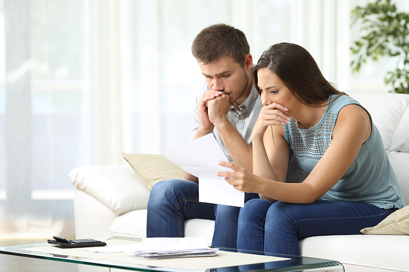 A man and woman sit on a couch looking concerned while reading a document together, with a remote control on the table in front of them.