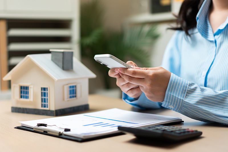 A person in business attire uses a smartphone at a desk with a model house, documents, pen, and calculator.
