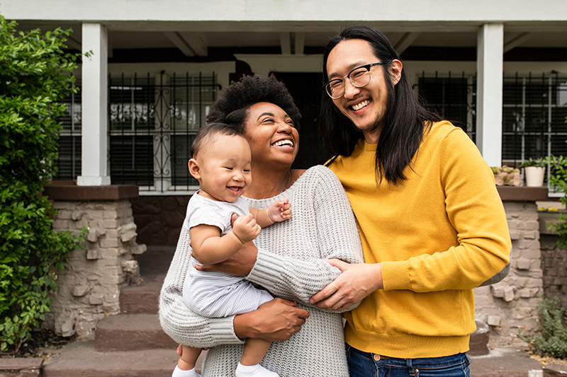 A smiling couple stands outside a house, with one person holding a laughing baby.