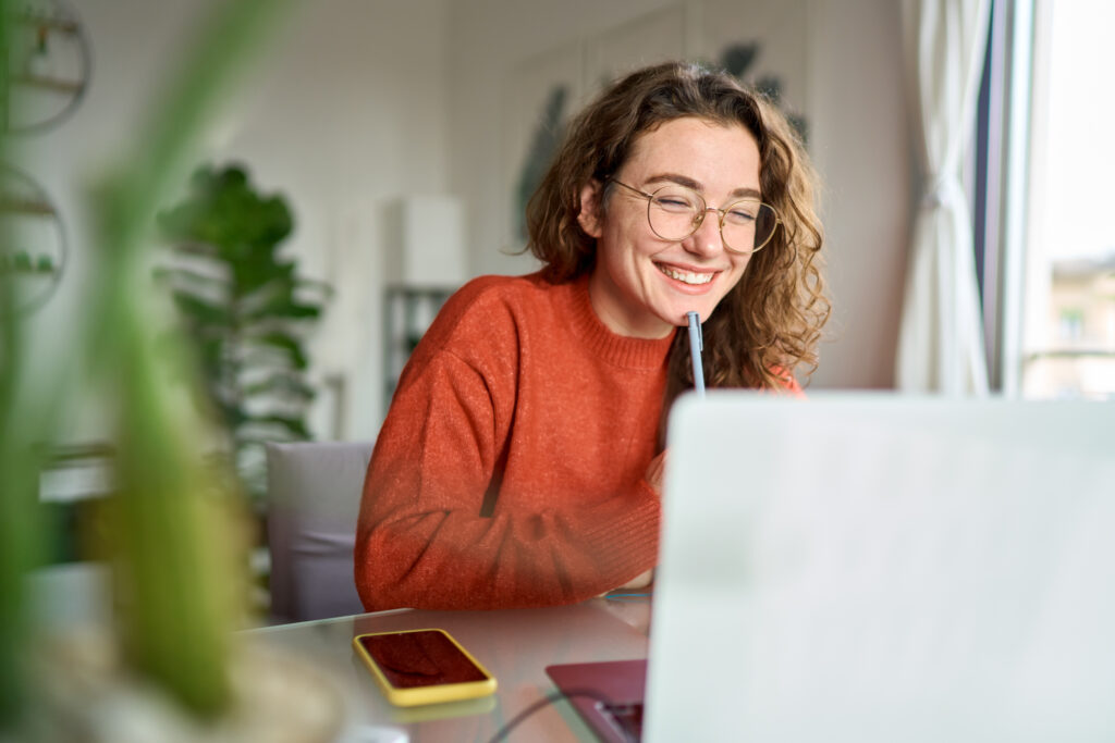 Woman wearing glasses and an orange sweater smiles while looking at a laptop, sitting at a desk with a phone and a plant visible in a bright room.