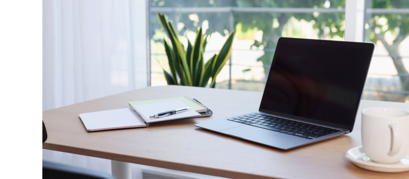 A laptop, notepad, pen, smartphone, and coffee cup are on a wooden desk near a window with a plant and greenery visible outside.