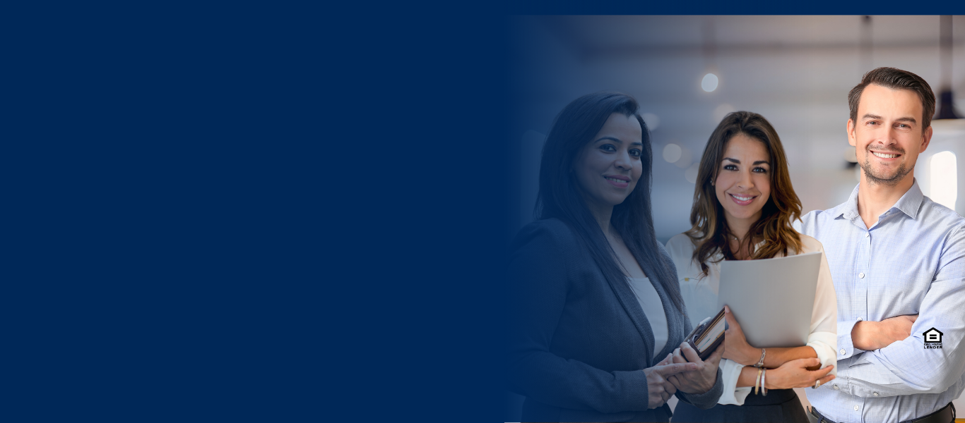 Three professionally dressed people, two women and one man, stand together smiling in an office setting with a large blue area on the left side of the image.