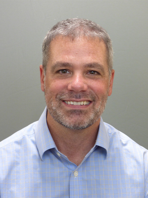 A man with short gray hair and a beard smiles at the camera, wearing a light blue, striped button-up shirt, against a plain gray background.