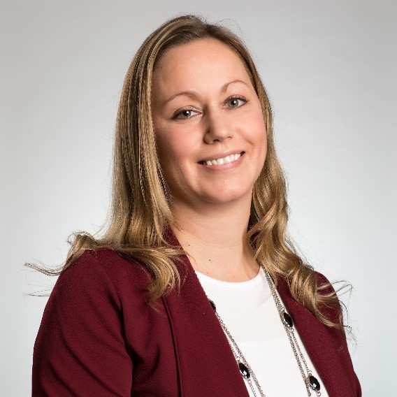A woman with long light brown hair, wearing a maroon blazer over a white top, smiles at the camera against a plain light background.